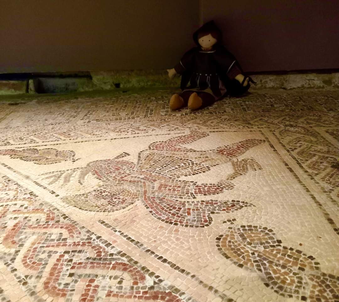 Our mascot 'Winter' gazing out over the mosaic that inspired his creation. He holds a leafless branch and a hare and wears a thick hooded cloak in the local style, called a birrus britannicus. #Mosaics