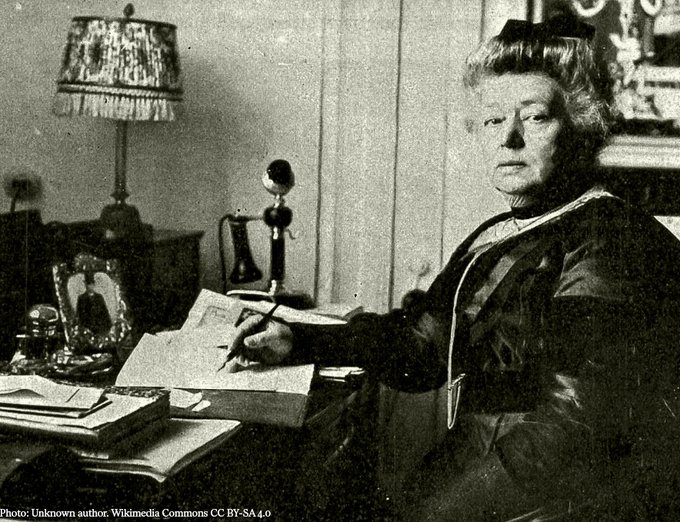 Bertha von Suttner sits at a desk with a pen in her hand, appearing to write a letter. 