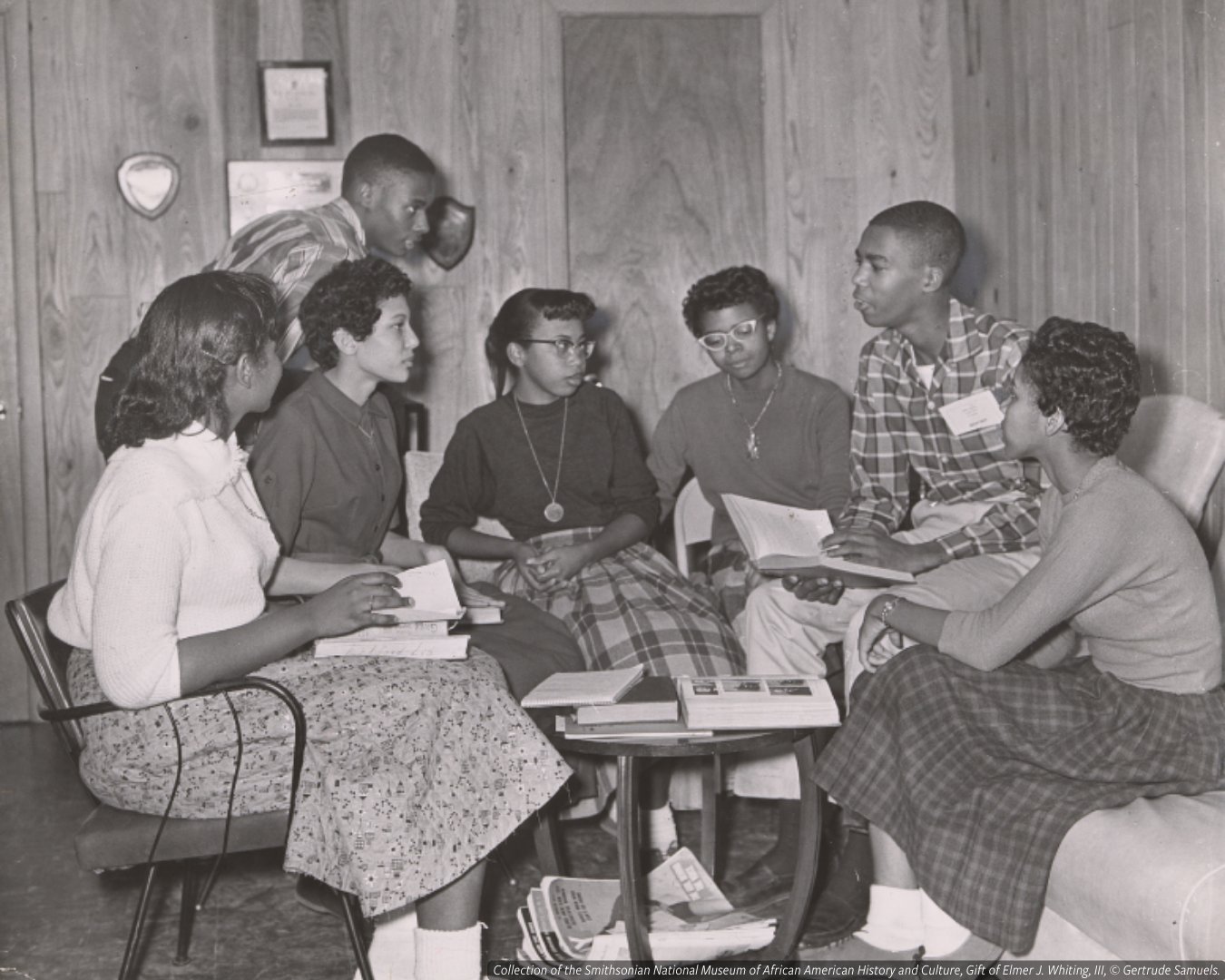 This black-and-white photograph depicts seven of the Little Rock Nine, from left to right, Melba Patillo, Carlotta Walls, Jefferson Tomas, Elizabeth Eckford, Thelma Mothershed, Terrence Roberts and Gloria Ray, meeting at the home of Daisy Bates, a local NAACP leader. The seven are gathered around a table with books. 