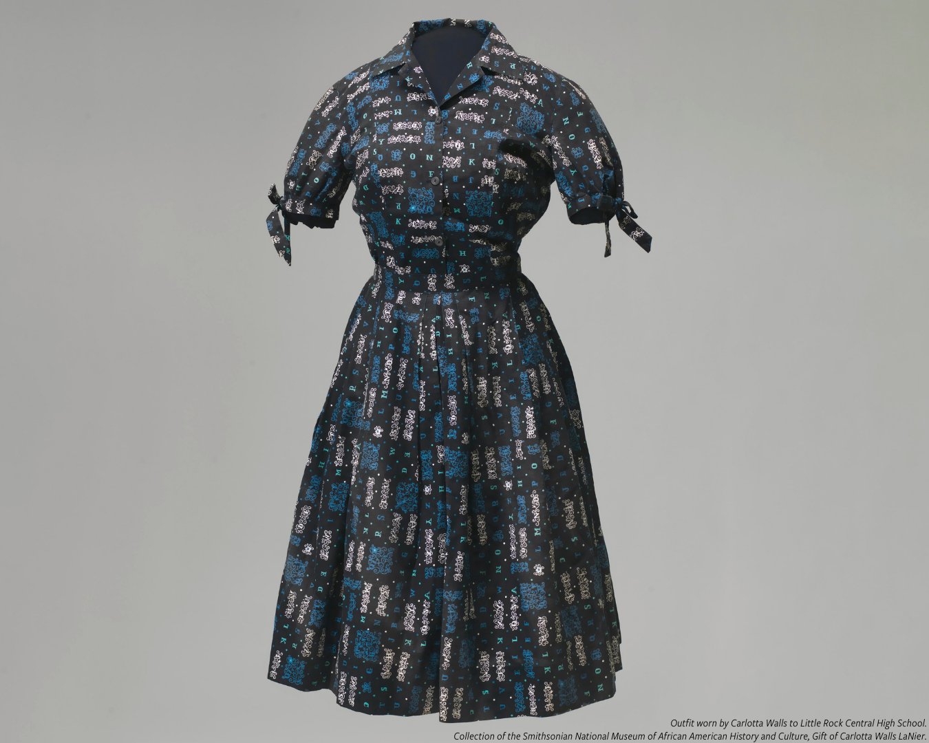 A color photo of the outfit worn by Carlotta Walls LaNier during the "Little Rock Crisis" of 1957 at Little Rock Central High School. The outfit consists of a skirt and blouse in a matching alphabetical print of blue, teal, and white on a black background. The blouse fastens with small black buttons, has a pointed collar and short sleeves that tie into bows at the cuffs. The skirt is full and has several pleats at the waist.