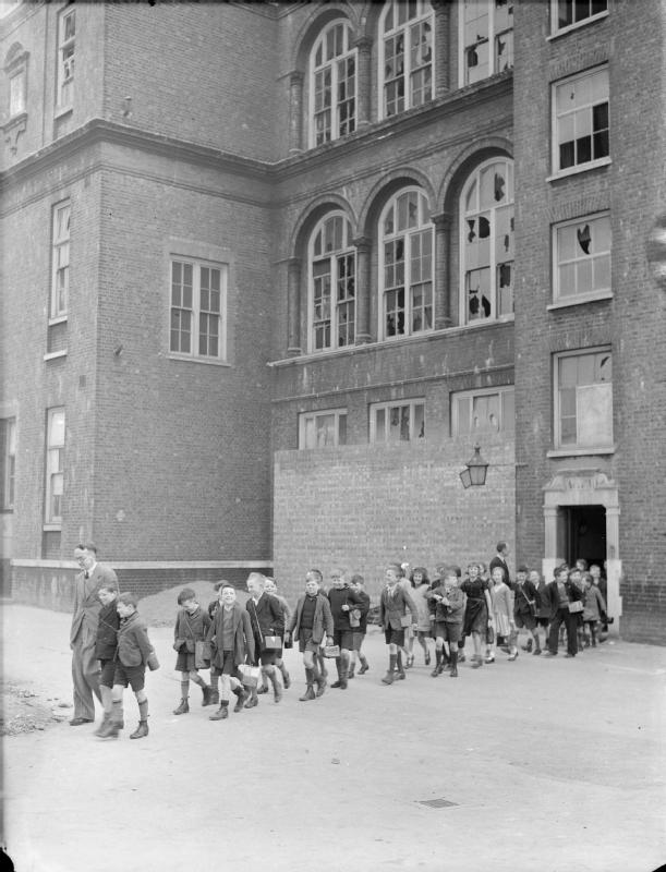 London Schools in Wartime- School Life in London, England, 1941 A teacher leads a large group of children out of the entrance of Old Woolwich Road School in Greenwich, London. The children are crossing the playground in a line two abrest and all carry their gas masks. The school