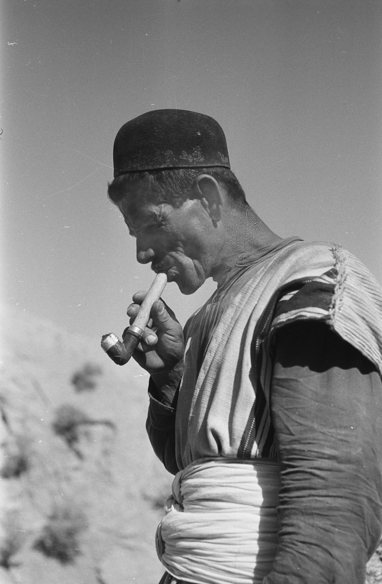 Portrait of a man, standing, wearing a black felt hat, smoking a pipe. Iran, 1964.