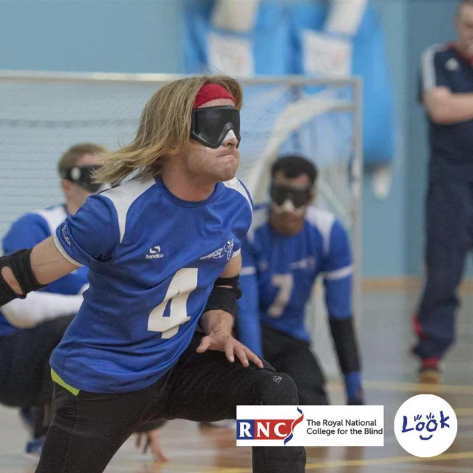 Image of mentor Warren kneeling down with a blue ball in his hand. He has a blindfold on and is wearing a blue sports top with the number 4 on it and black sports trousers, in the background are three other goalball players in the same kit and a goal.