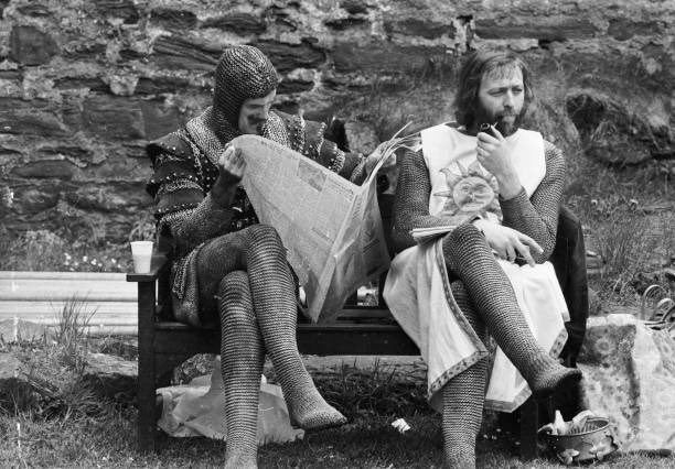 A chainmail-clad John Cleese reads a newspaper while Graham Chapman smokes a pipe on the set of “Monty Python and the Holy Grail,” a new medieval-themed comedy film that will come out next year.