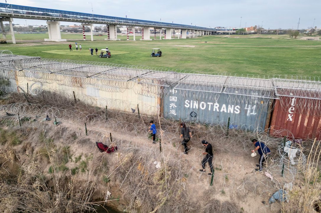 Migrants walk on the banks of the Rio Grande river to find a way to enter the United States as golfers play on the other side of the shipping container fence in Eagle Pass, Texas in March 2024 (Go Nakamura/Reuters)