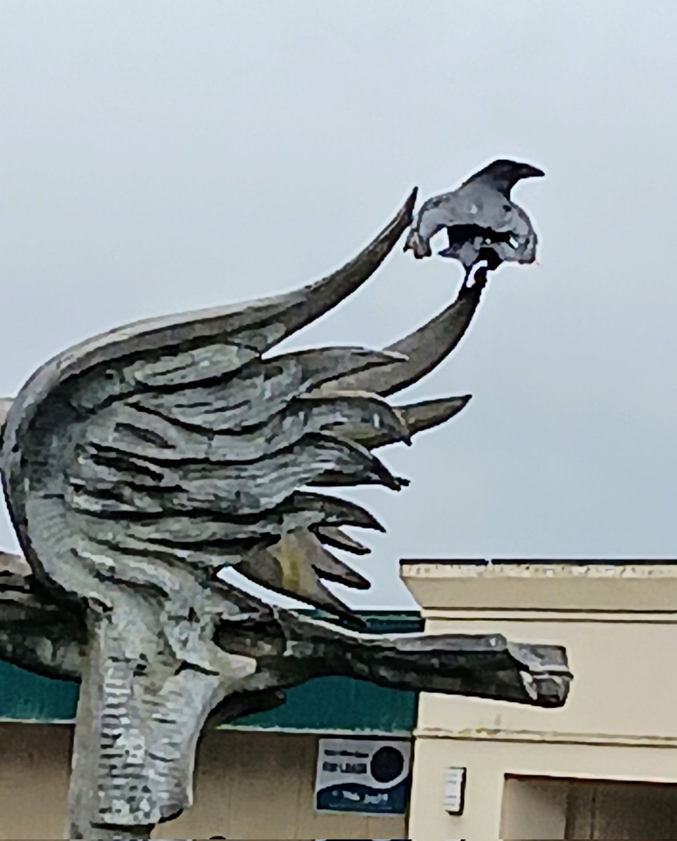 A raven showing off by posing on a concrete eagle statue on a windy day.