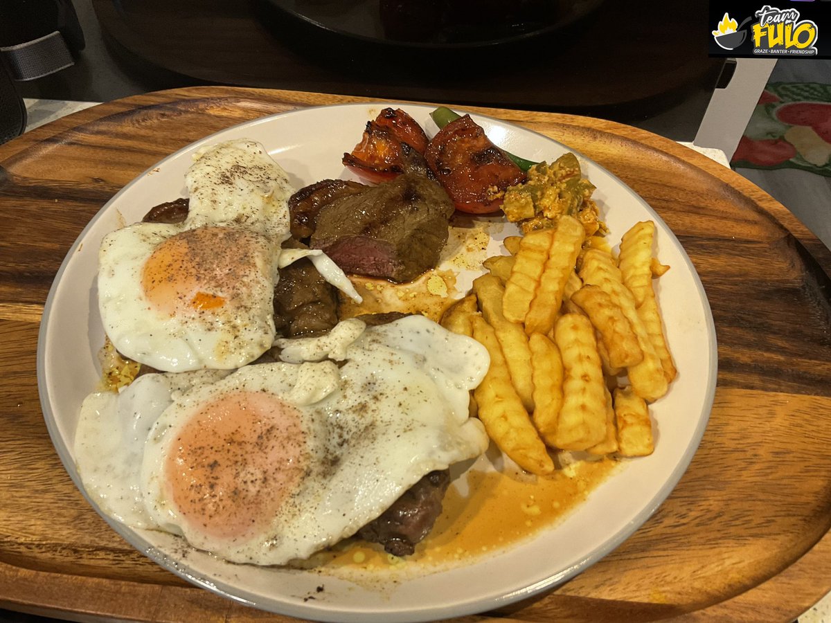 Two sunny-side up ,ooooozy,woooozy free range golden yolk eggs laid gently on top of a bible thick slab of juicy,succulent steak ,pan fried to medium with a pouring of its own juices. Grilled tomato,crinkle cut chips ,Sista Janet Buys’ Zimbabwean homemade fiery mango pickle & 2