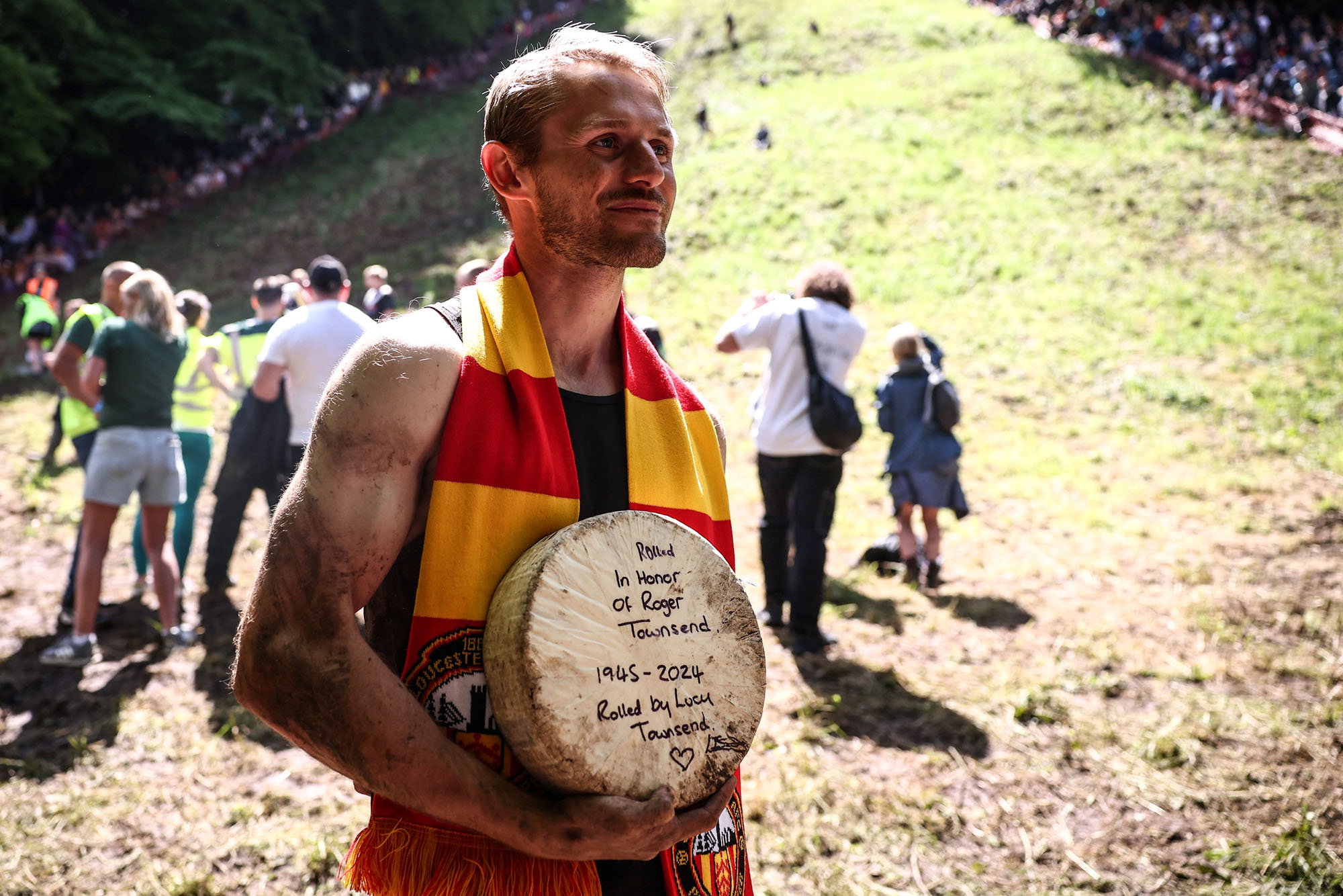 A muddy, but cheerful, man poses at the bottom of a steep hill, holding his prize, a round of cheese. Written on the round are the words "Rolled in honor of Roger Townsend, 1945-2024. Rolled by Lucy Townsend."