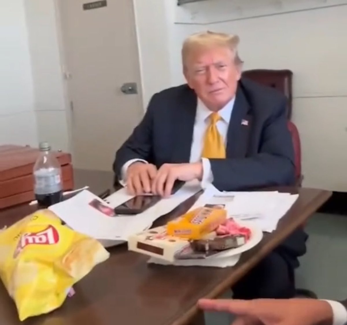 A picture of Trump from the courthouse surrounded by junk food: a bag of lays, a Diet Coke, and boxes of candy including Milk Duds, Whoppers, and others.