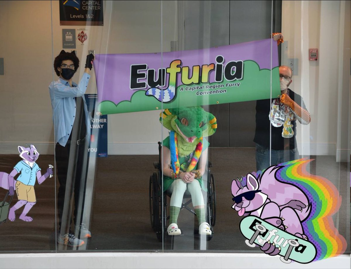 Two con attendees holding up a large Eufuria banner over a snake fursuiter seated in a manual wheelchair