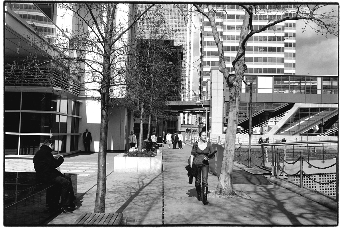 Figures in the city no. 204 Millwall Inner Dock, London, 2012 Silver gelatin print #London #city #CanaryWharf #streetphotography #woman #men #figures #blackandwhitephoto #monochrome #analogue #film #bnw #urbanlandscape #cityscape #towers #apartments #trees #shadows #sunlight
