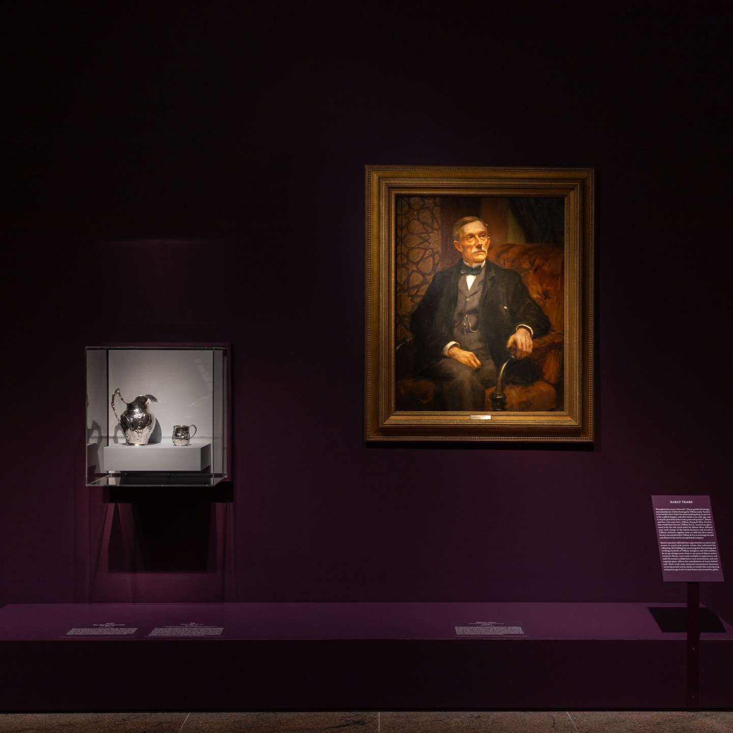 Portrait of a man displayed in a dark museum setting, beside a glass case containing silver objects. 