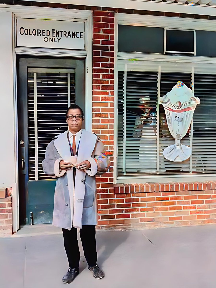 Writer James Baldwin stands outside an ice cream parlor with a "Colored Entrance Only" sign above the door. Behind him, a worker from the parlor peeks out through the window blinds. 

Durham, North Carolina, 1963.