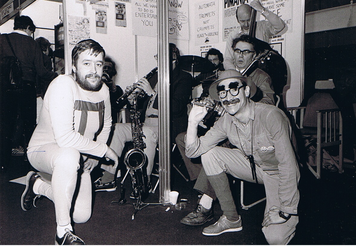 Christian Penfold and Mel Croucher of Automata at a trade show in the early-mid 80s. In protest against the violence in computer games, Automata had cleared its stand to make way for jazz band. Note the posters behind the band: "Trumpets Not Crumpets, Peace Not Violence"