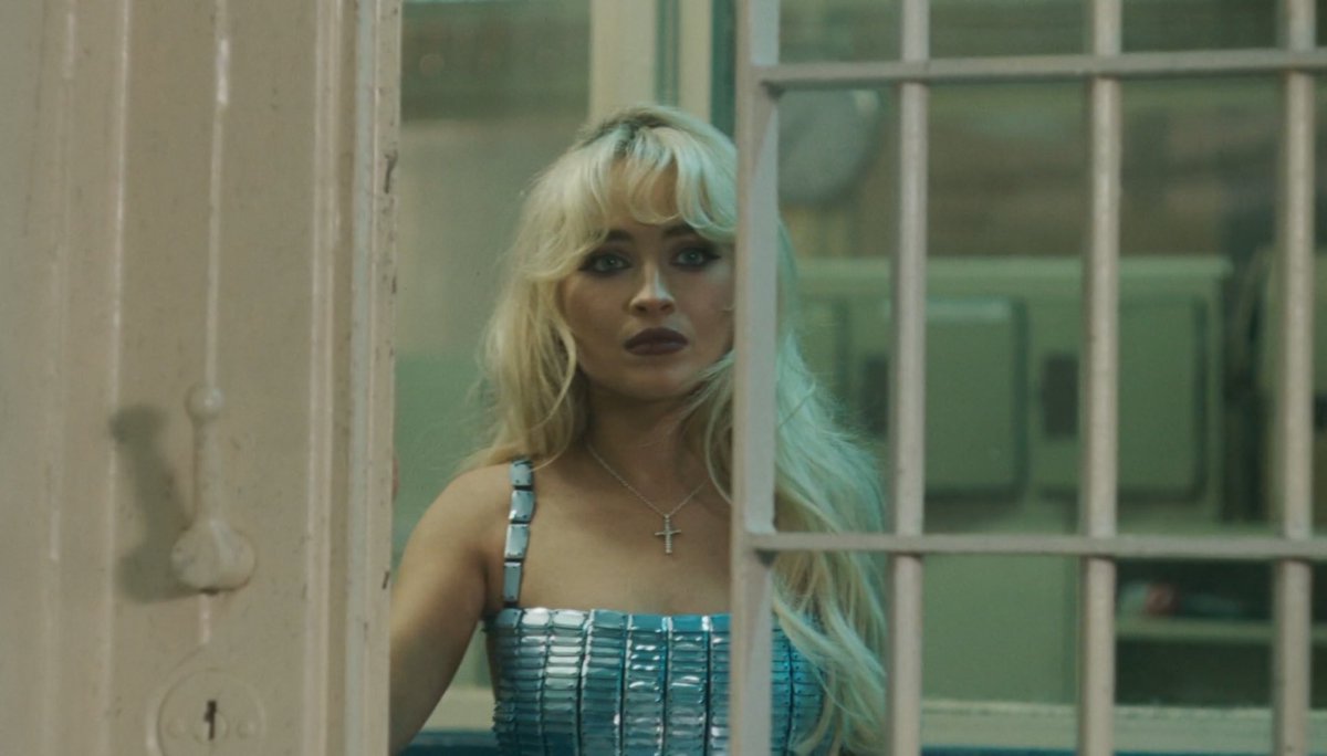 A girl with heavy makeup and long blonde hair wearing a blue dress and a silver cross stands behind the closing gate of a white jail cell.