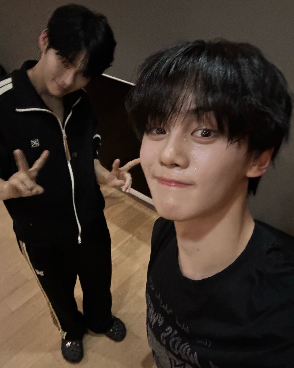 Gyuvun's tweet image. This photo was taken during a dance practice with Hanbin hyung a while back. I’m up front, smiling with a bit of sweat on my face, while Hanbin hyung is in the background flashing a peace sign. The dimly lit studio captures the intensity and focus of our practice sessions.