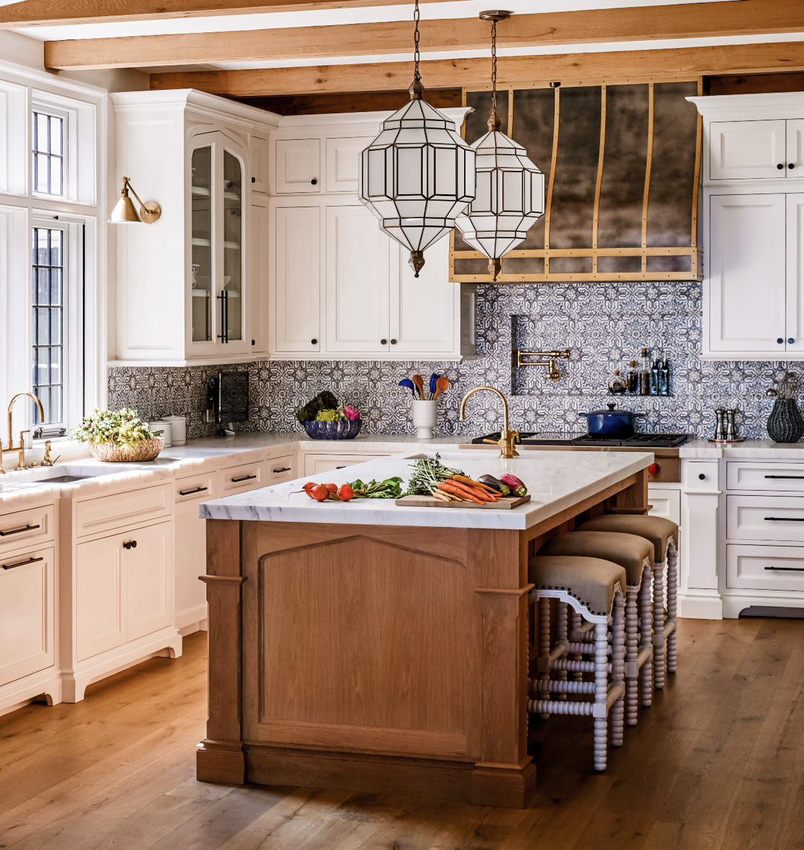 The bright kitchen in our Hilltop English Country home combines both rustic and refined finishes, including its metal range hood, painted white cabinetry, saturated blue-gray backsplash, and natural wood island &amp; beamwork.⁣ #kitchendesign #luxuryhome #architects #designdetails