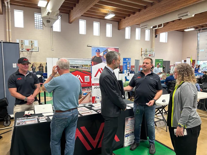 People engaging in conversation at a job fair inside a gymnasium with various promotional stands. There's a noticeable "JobConnect" banner and a sign reading "Hire Heroes USA".