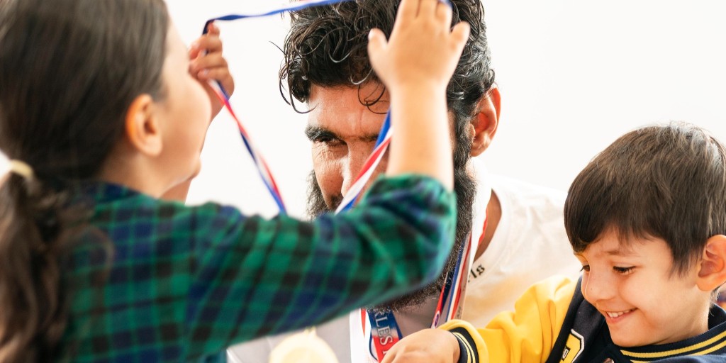 Wali Noori - a blind afghan refugee - is shown wearing a number of medals with two children - a boy and a girl - alongside him. He holds the young boy, who is playing with the medals, whilst the girl places a further medal over his head.