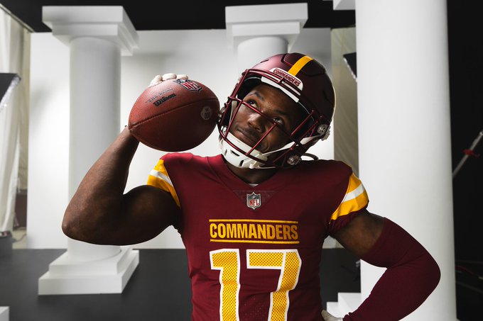 Photo of Terry McLaurin. He is wearing a burgundy helmet, jersey and sleeve on his left arm. He is holding a football in his right hand and holding it up to his face. He is smiling and looking up towards the sky. In the background of the photo are white columns. 
