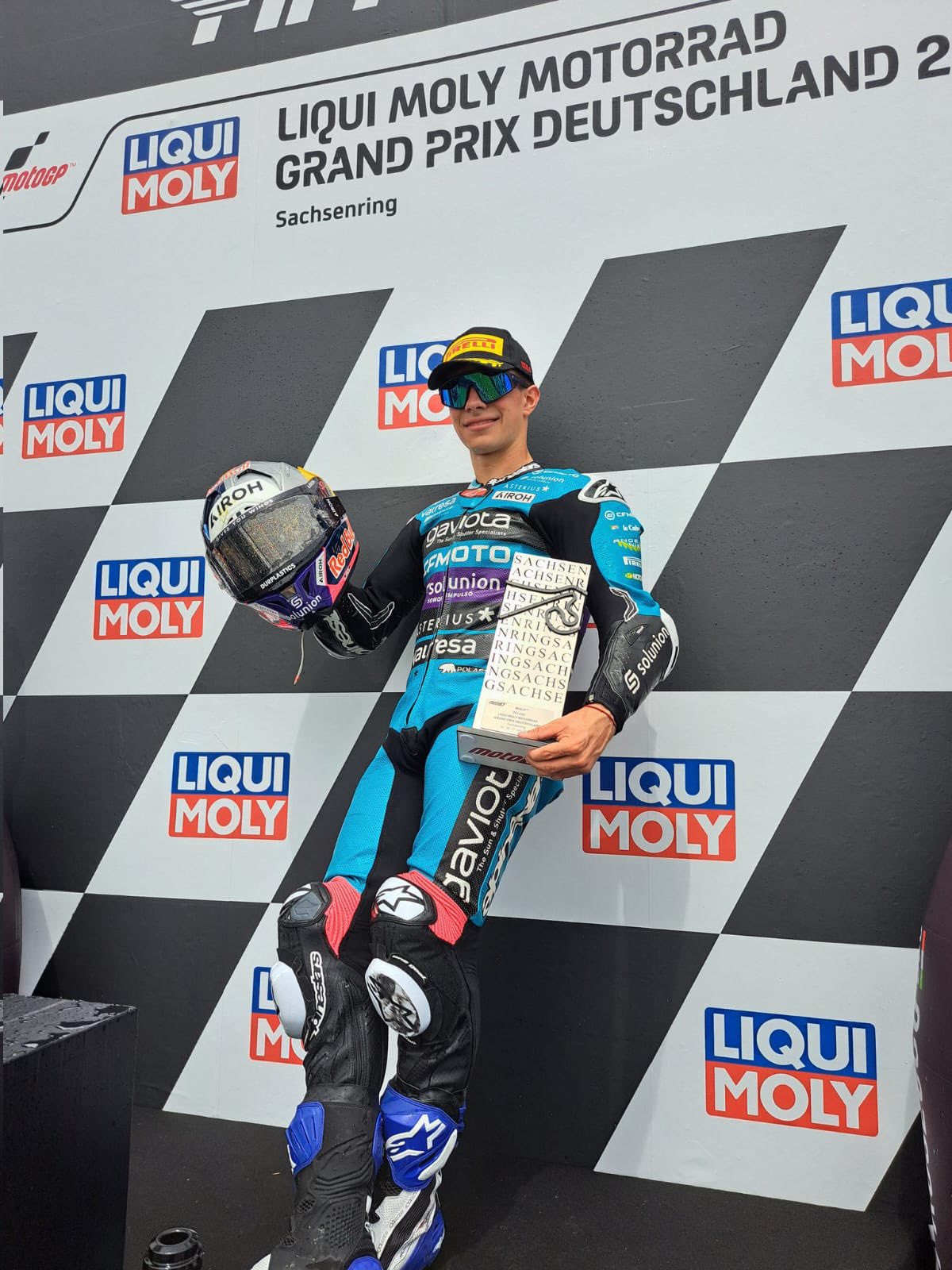 📸: @AsparTeam. David Alonso con casco en su mano derecha y en la izquierda un trofeo. Usa uniforme azul con negro, gorra y lentes oscuros. Apoya su espalda en un backing con logos de la competencia