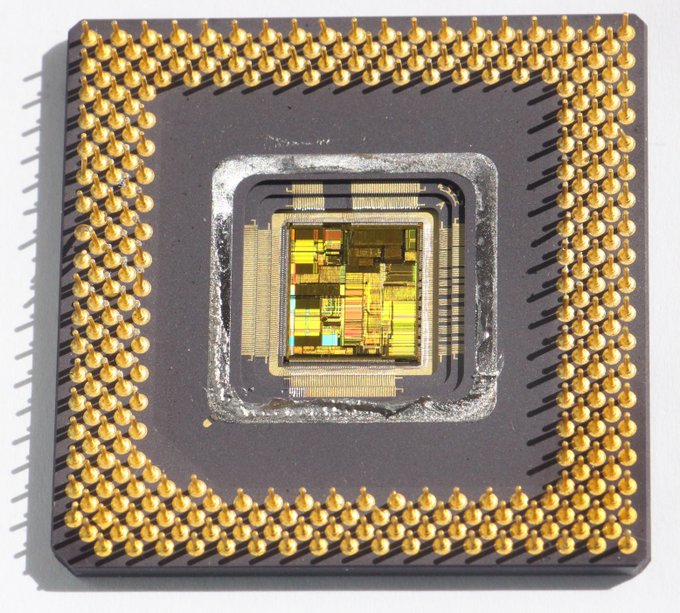 A photo of the Pentium processor in its ceramic package. The package has many golden pins sticking out of it. The silicon chip itself is exposed in the middle. The patterns on the chip are visible in shimmering colors.