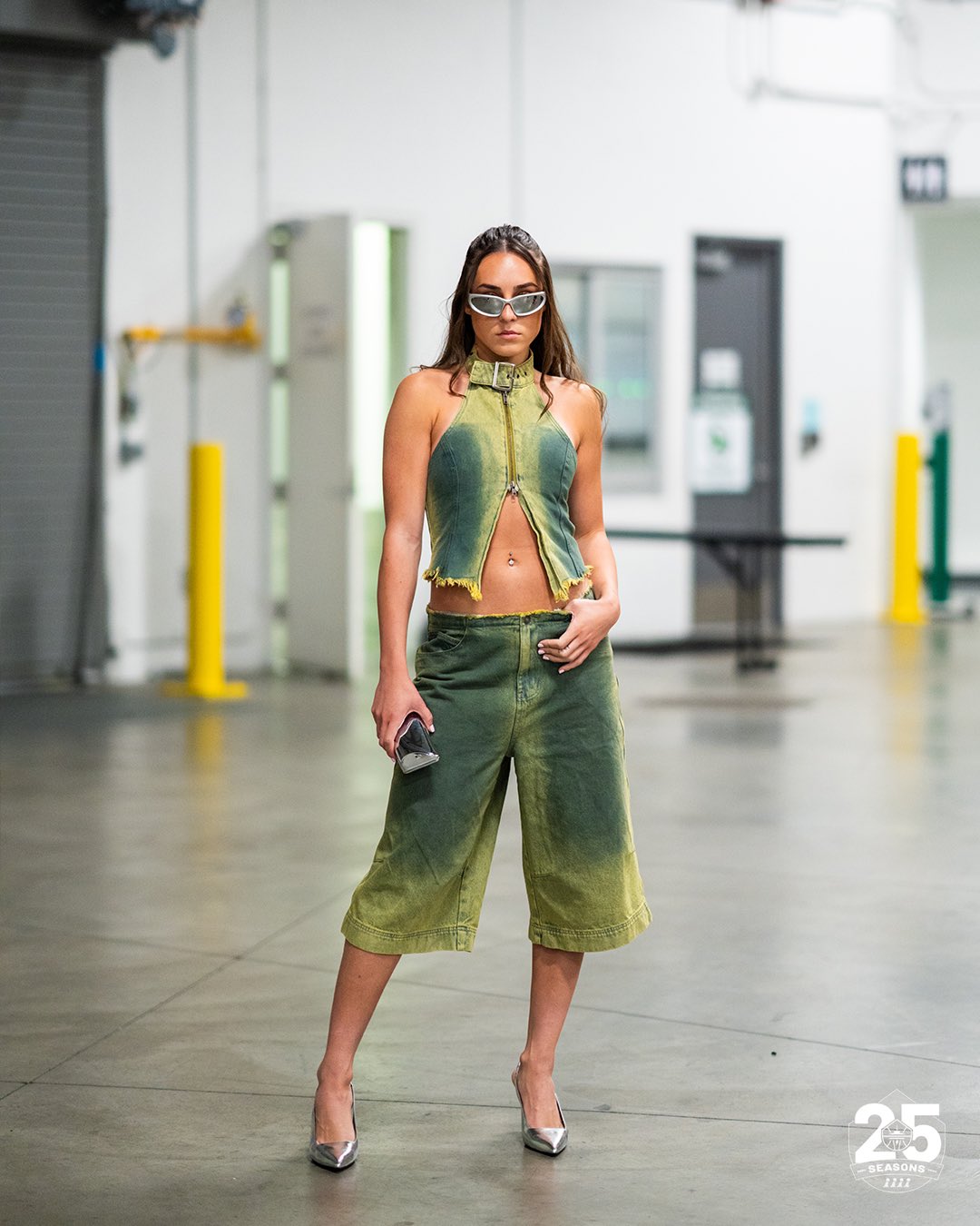 Nika Mühl posing in the loading dock in Climate Pledge Arena wearing a matching distressed denim set with silver sunglasses and heels