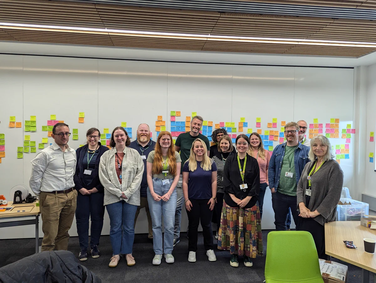 A friendly and smiley group of colleagues who have just completed their UX Training. They are standing in front of a white wall with lots of colourful post-it notes on. 