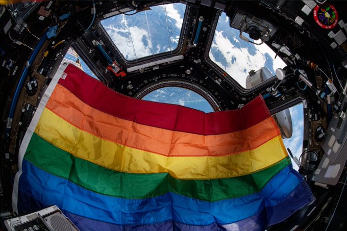 A rainbow-colored Pride flag floats in the cupola aboard the International Space Station. The flag is colored in stripes of red, orange, yellow, green, blue, and purple, and takes up most of the image. Behind it, though, the windows of the cupola can be seen looking down at Earth. Credit: NASA/Raja Chari