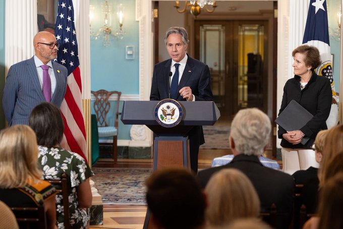 Secretary Blinken speaks while standing at a lectern, while a family member of former Secretary Albright and a family member of former Secretary Powell stand at on both sides.