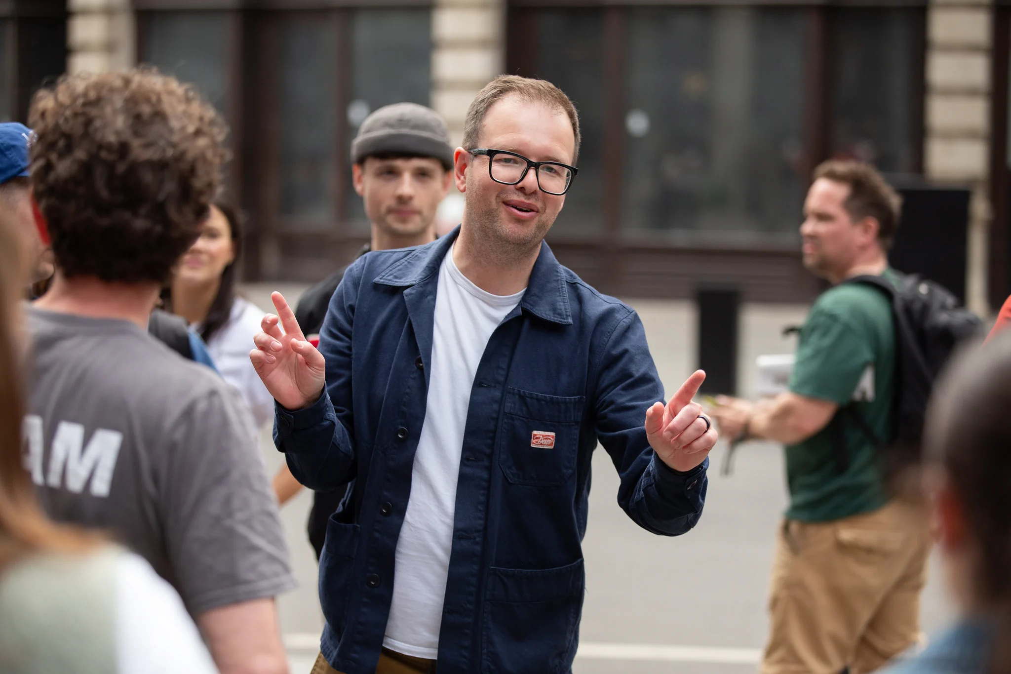 A man wearing glasses and a blue jacket gestures with both hands while talking to a group of people outdoors. Other individuals are visible around him, some paying attention, in a city setting with blurred buildings in the background.