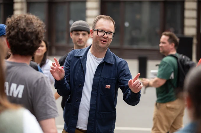 A man wearing glasses and a blue jacket gestures with both hands while talking to a group of people outdoors. Other individuals are visible around him, some paying attention, in a city setting with blurred buildings in the background.