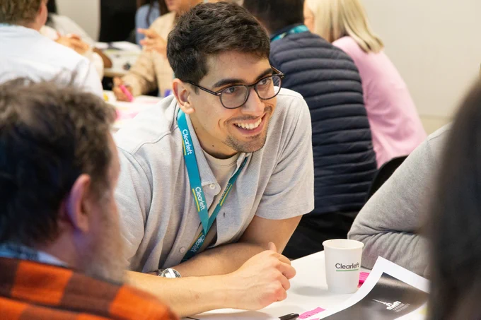 A man with dark hair and glasses, wearing a gray shirt and a blue lanyard, is smiling and engaging in conversation with others around a table in a brightly lit room. The table has papers, a coffee cup, and other materials on it.
