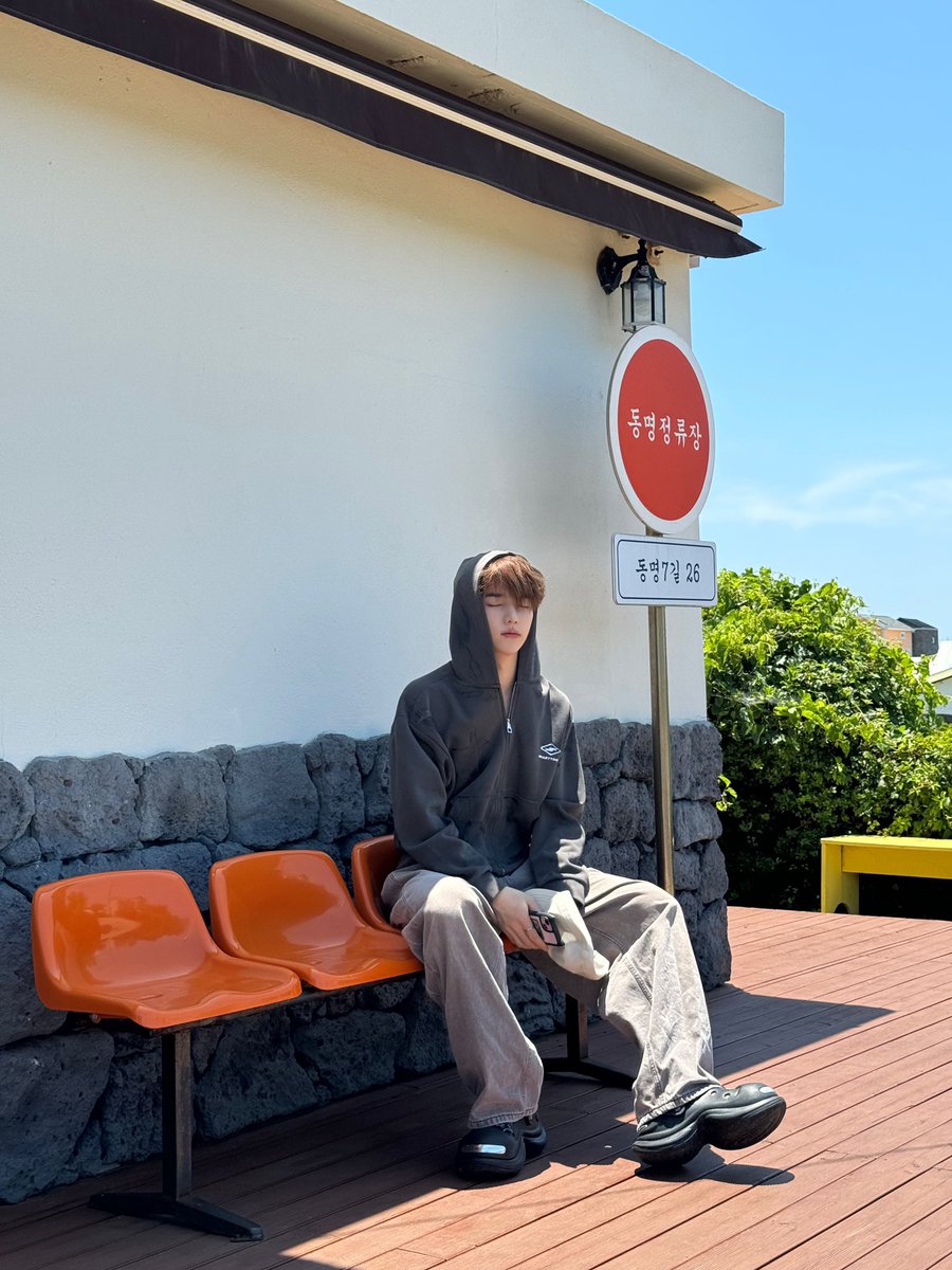 Sitting on bright orange plastic chairs, I feel the coolness of the gray stone wall behind me. The sun is warm, but my gray hoodie and loose pants keep me comfortable. I hold my beige cap loosely, glancing around at the serene setting.