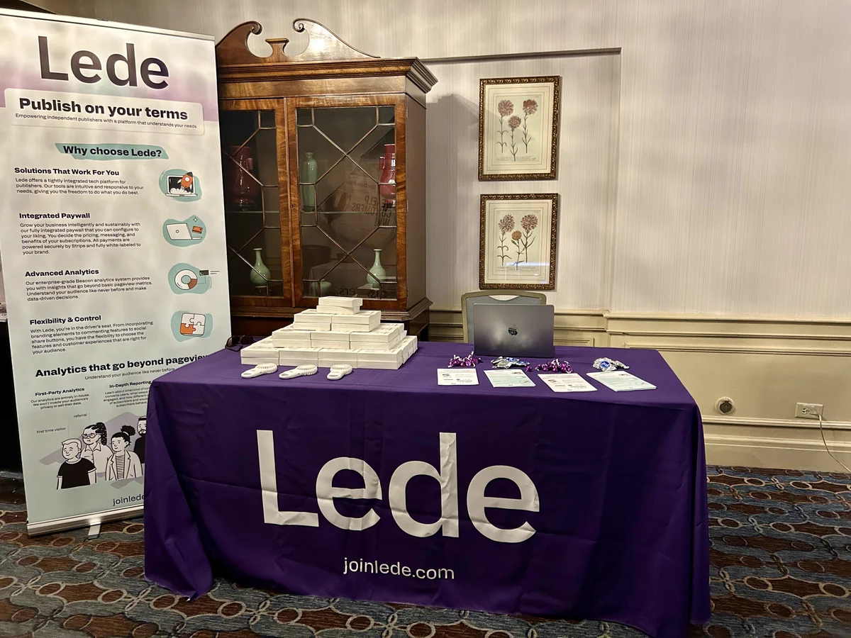 Lede's vendor table at the AAN conference is covered in a bright purple tablecloth that reads "Lede." On the left side of the table is our swag - a white handheld mini fan. Three fans are out of their boxes and behind those samples is a pyramid of fan boxes. On the right side of the table is an open laptop, some handouts, and some purple and silver candy. To the left of the table is a popup banner with info about Lede and some whimsical graphics, including a spaceship launching out of a computer. 