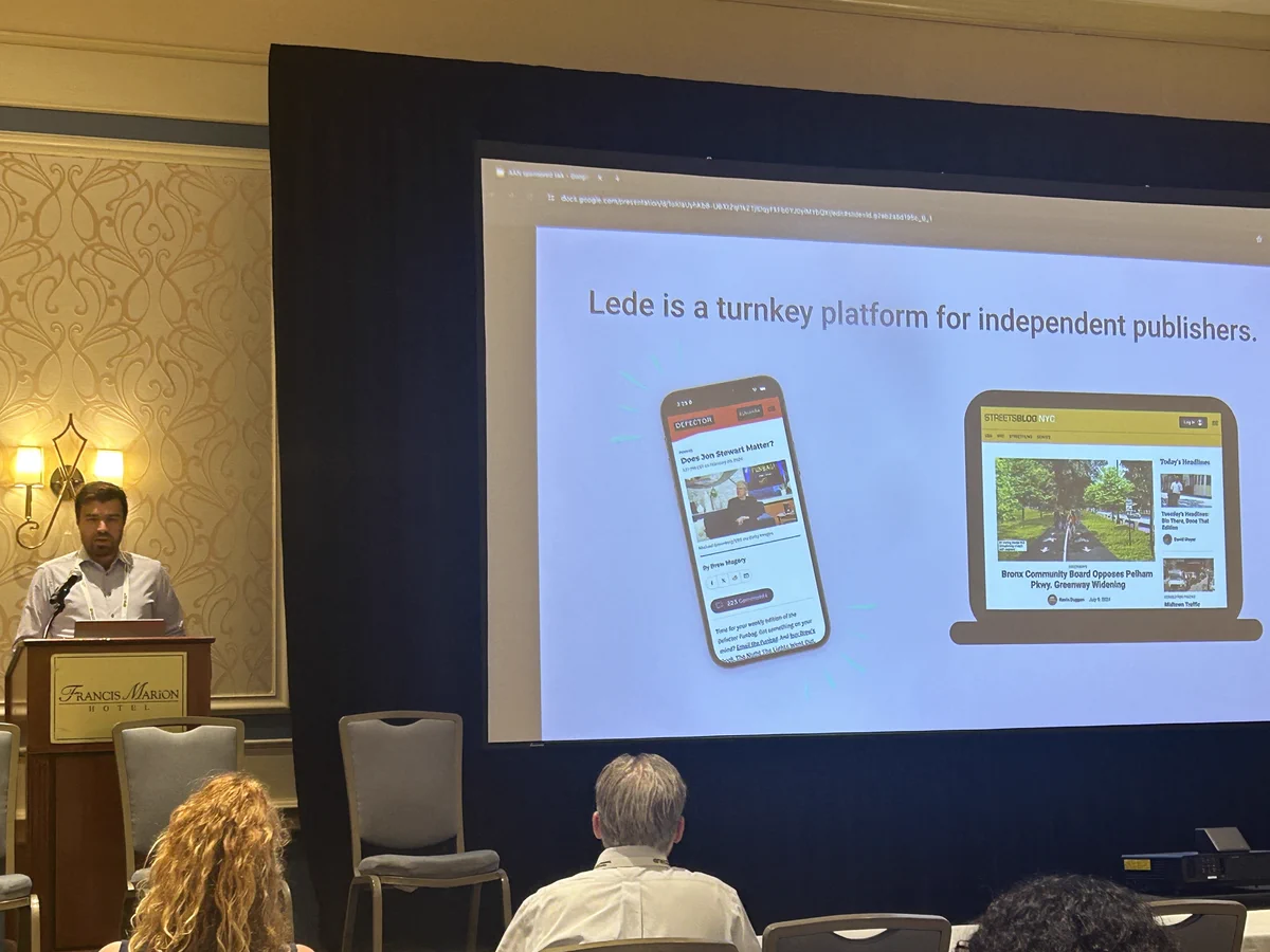 Lede CEO Matt Johnson standing at a lecturn in a hotel ballroom talking about Lede. To his right is a large screen with the heading "Lede is a turnkey platform for independent publishers" and two images. The image on the left is a mockup of a mobile device featuring the homepage of Defector, a Lede publisher. The other image is a mockup of a laptop featuring the home page of Streetsblog, another Lede publisher. Only the first row of people in the ballroom is visible.