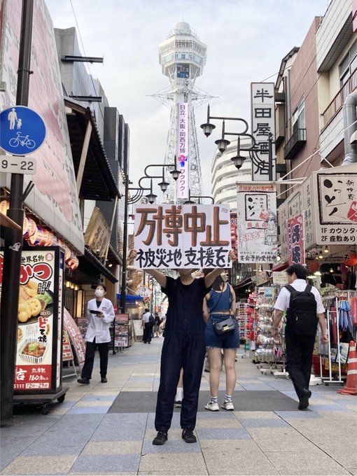 大阪は通天閣前に立つ人。手には「万博中止 被災地支援を！」と書かれたプラカード。