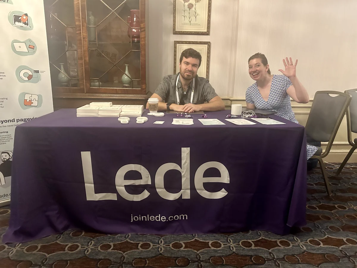 Lede CEO Matt Johnson and Lede principal software lead Christina Deemer sit at a conference booth looking at the camera. Christina is waving at the camera with her left hand. The table is covered in a purple linen with that says Lede in white letters and Lede's website address, joinlede.com. 