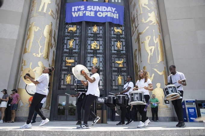 a drumline performs under a banner reading "It's Sunday. We're open!" in front of the Brooklyn library