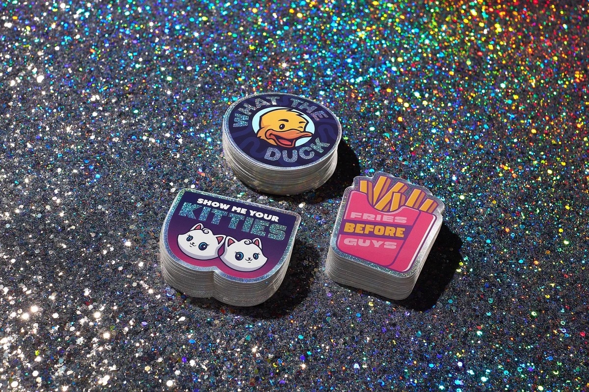 Three stacks of glitter stickers sitting on a glitter-filled table top. One sticker is of a pink and yellow order of French fries that says “fries before guys.” The second stickers is a circle with a cartoon duck and text around the edge that says “what the duck.” The third sticker shows two white cartoon cats with the text “show me your kitties” above them.