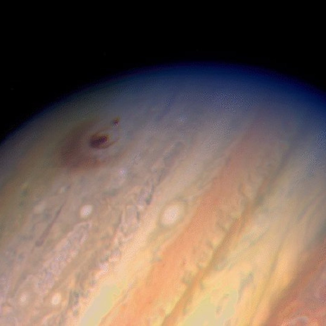 A portion of the planet Jupiter is seen against black space. Jupiter's atmospheric bands are seen in cloudy stripes of orange, cream, and brown. A faint, black impact scar from comet fragments is visible near the upper left of the planet.