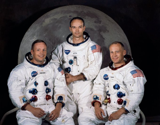 Neil Armstrong, Michael Collins, and Buzz Aldrin in white Apollo spacesuits pose for a portrait in front of a picture of the Moon