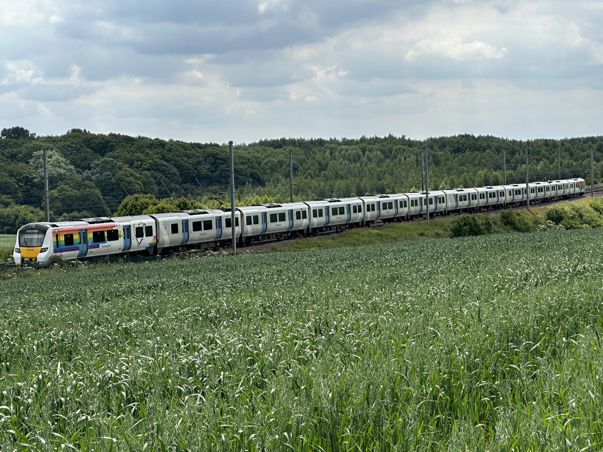 A Thameslink train with a rainbow design on it, travelling on a train track with fields, trees and greenery either side.