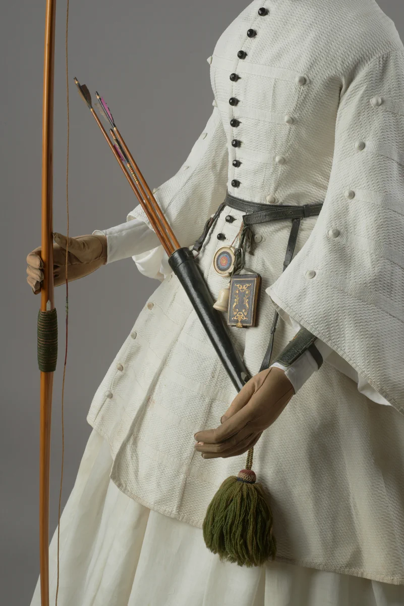 A close-up of mannequin dressed in a traditional archer costume, featuring a white tunic, full sleeves, and a layered skirt. The mannequin is accessorized with a brown quiver, holding arrows, attached to a detailed belt with a round, ornamental buckle.  