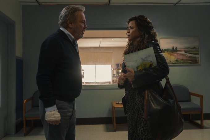 Billy Crystal and Rosie Perez look towards each other, worried, in a doctor’s office setting. Billy Crystal's hand is bandaged and Rosie Perez holds an unseen painting in her arms.