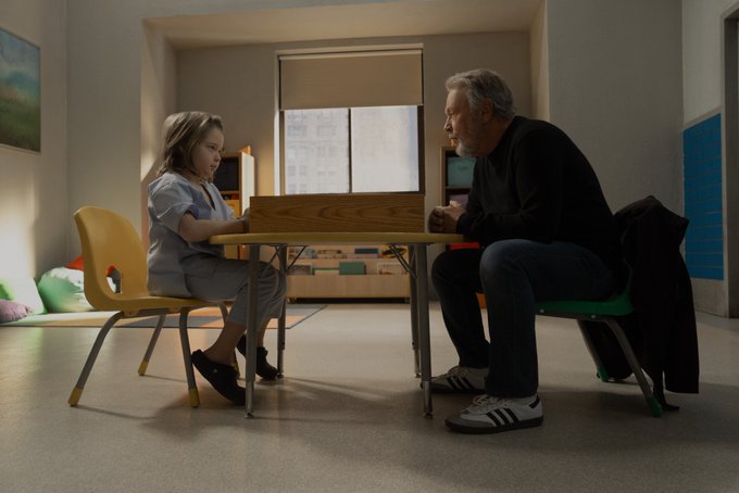 Billy Crystal sits across from a little boy in hospital scrubs. They are sitting at a table for children in a hospital playroom.