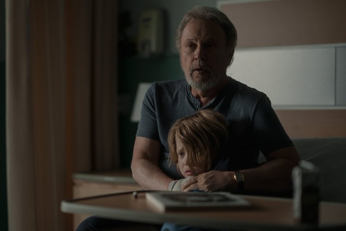 Billy Crystal holds on to a scared little boy as they both sit on a hospital bed looking forward.