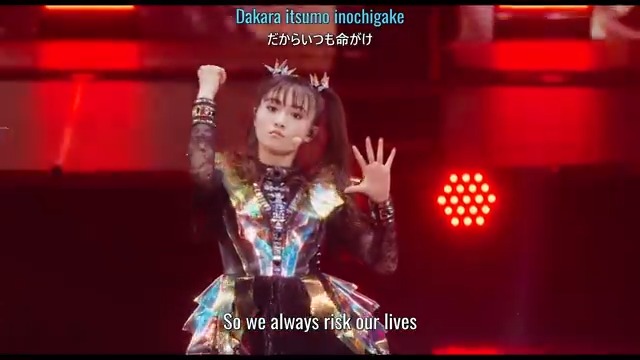 A BABYMETAL performer in a metallic black and gold outfit performs on stage, with on-screen subtitles reading 'Dakara itsumo inochigake' and 'So we always risk our lives.'