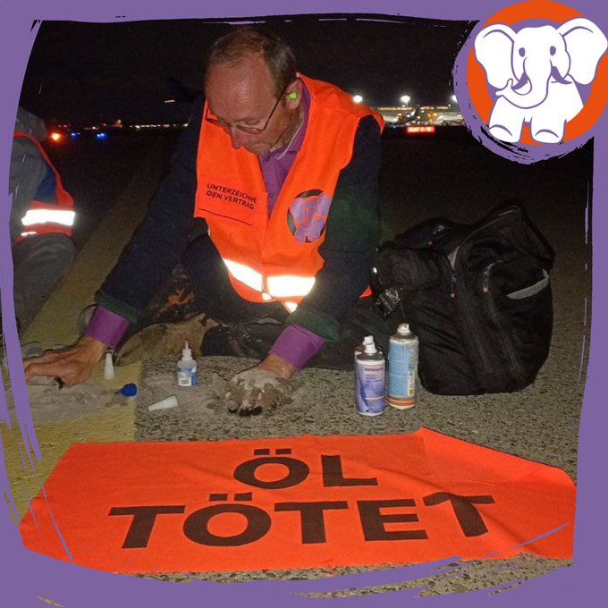 York sitzt auf einem Rollfeld, eine Hand auf dem Asphalt geklebt. Vor ihm liegt ein Banner mit dem Spruch „Öl Tötet“ 