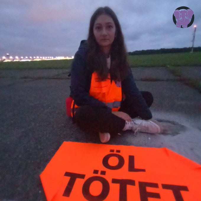 Emma sitzt auf einem Rollfeld vor ihr ein Organges Banner mit dem Spruch „Öl Tötet“ 